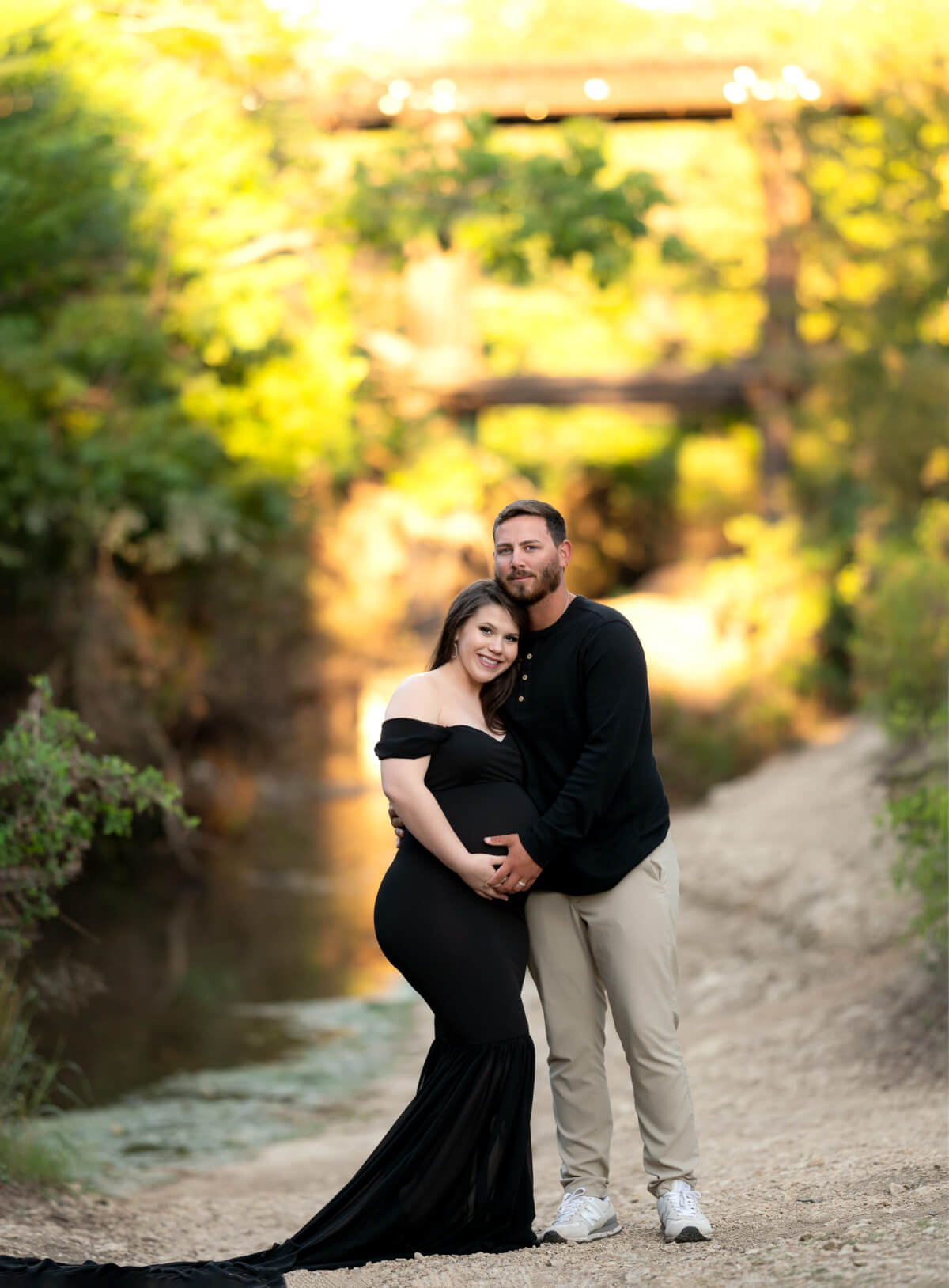 A man and a pregnant woman stand in front of a bridge over Brushy creek. They are looking toward the camera, and each have a hand on her stomach.