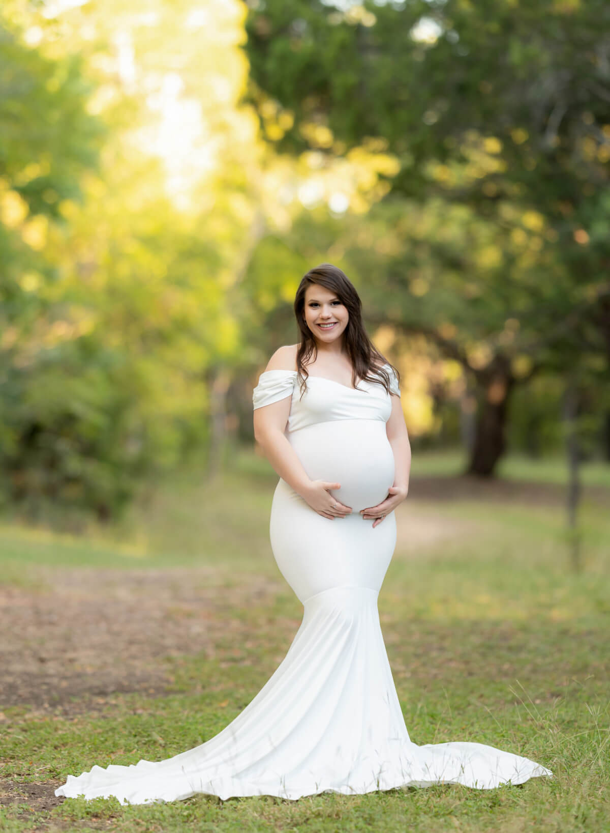 A pregnant woman stands in a path surrounded by trees. She is wearing a long, white maternity gown and cradling her baby bump. 