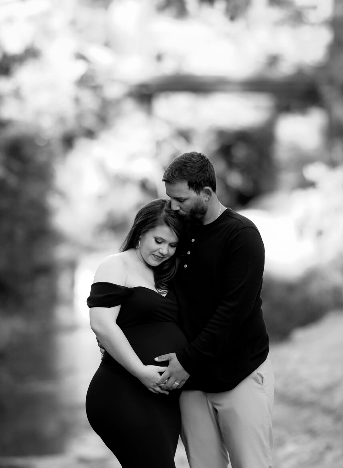 A black and white photo showing a man and woman in front of a wooded area in Brushy Creek park. The man is kissing the woman on the forehead, and they both have a hand on her pregnant stomach.