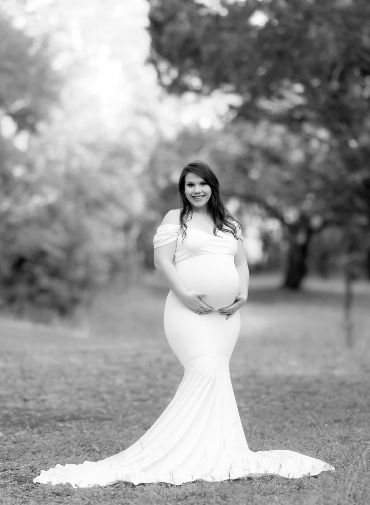 A black and white maternity photo featuring a woman in a long, white gown in a wooded grove. She is cradling her baby bump.
