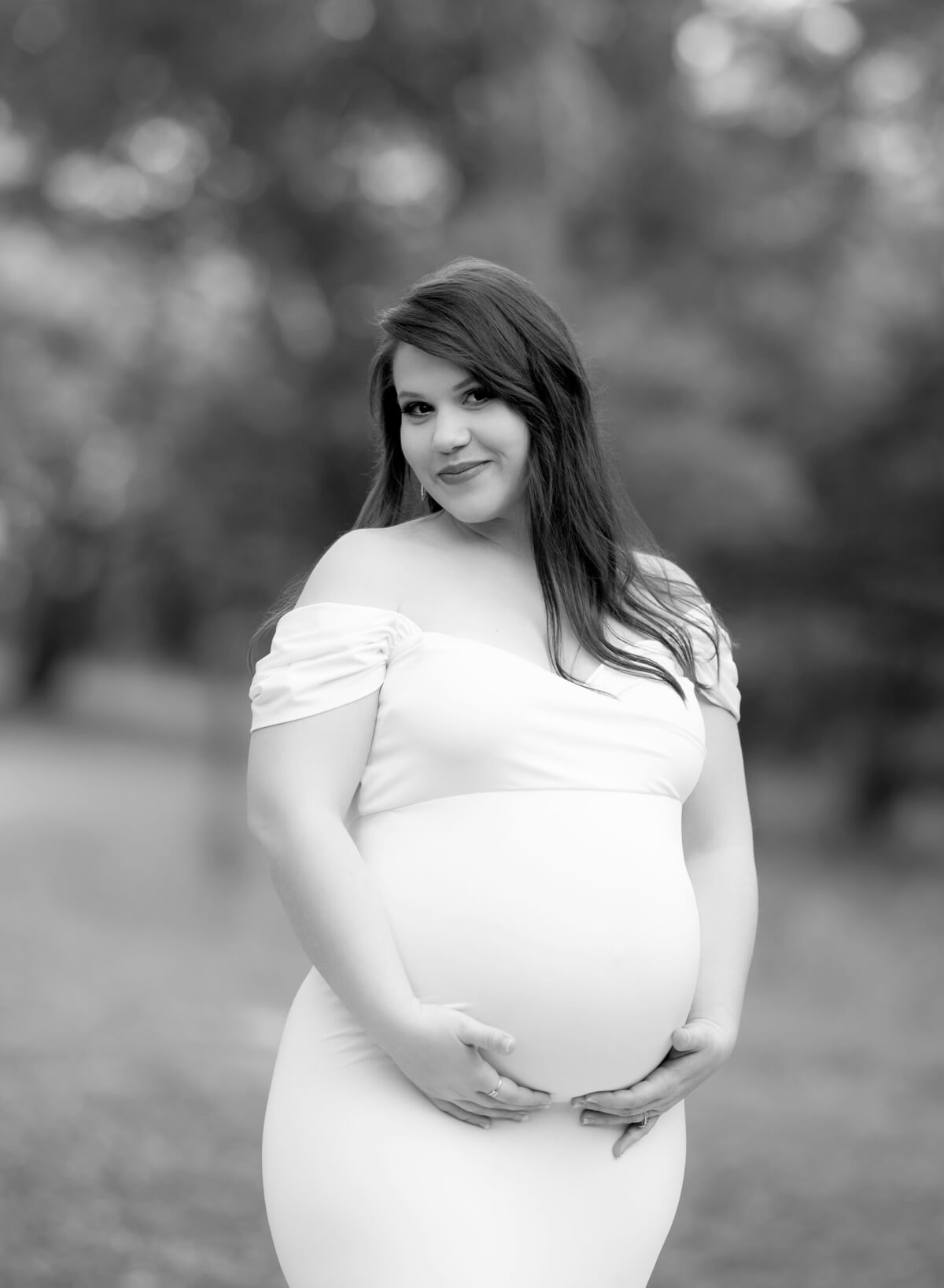 A black and white maternity photo featuring a pregnant woman holding her tummy in a wooded area.