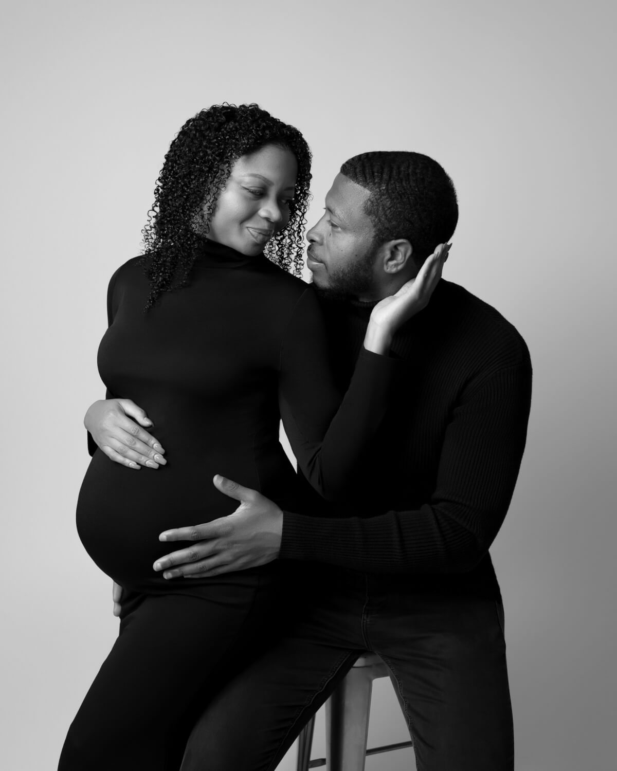 A man sits on a stool and holds his pregnant wife in his lap. 