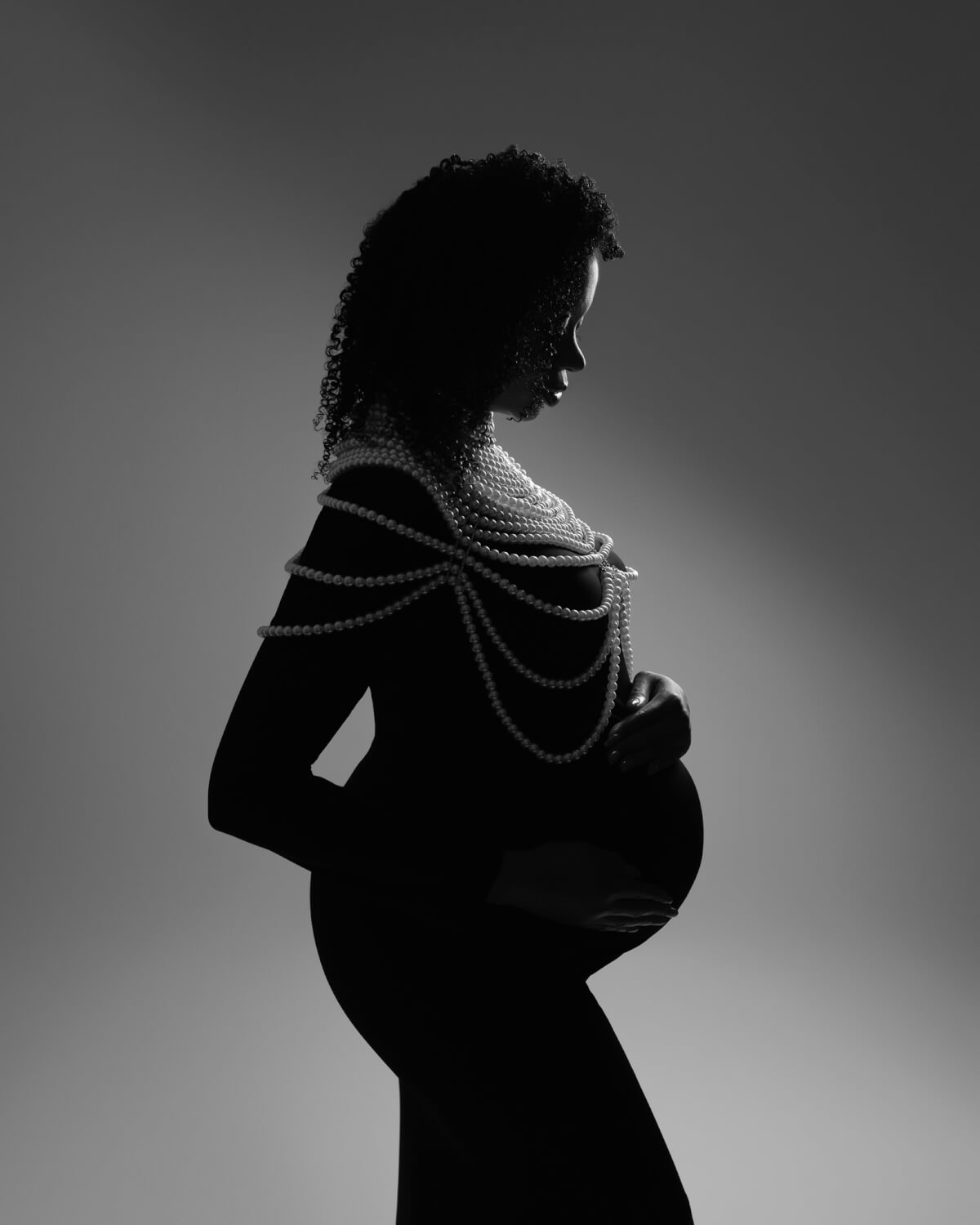 A richly shadowed side portrait of a pregnant woman in a black gown holding her stomach. Her shoulders are draped in pearls. 