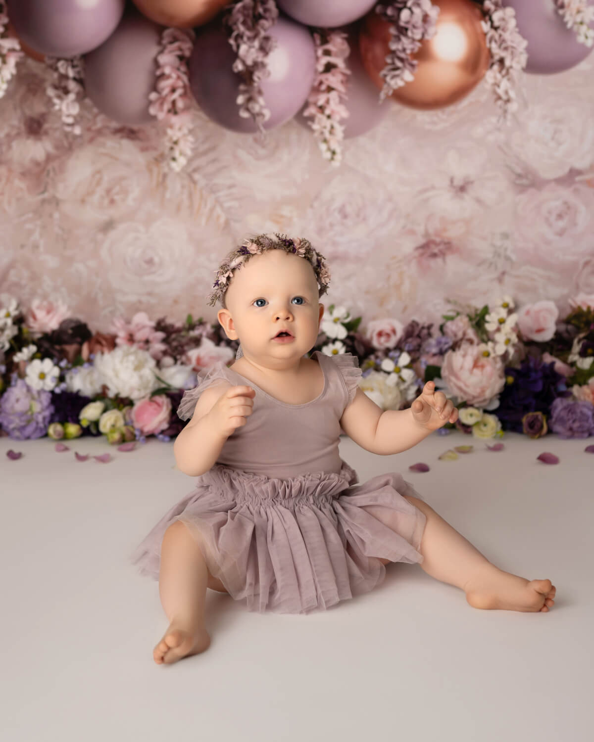 A one-year-old baby sits in front of balloons and flowers. She is wearing a floral headband and a lavender dress