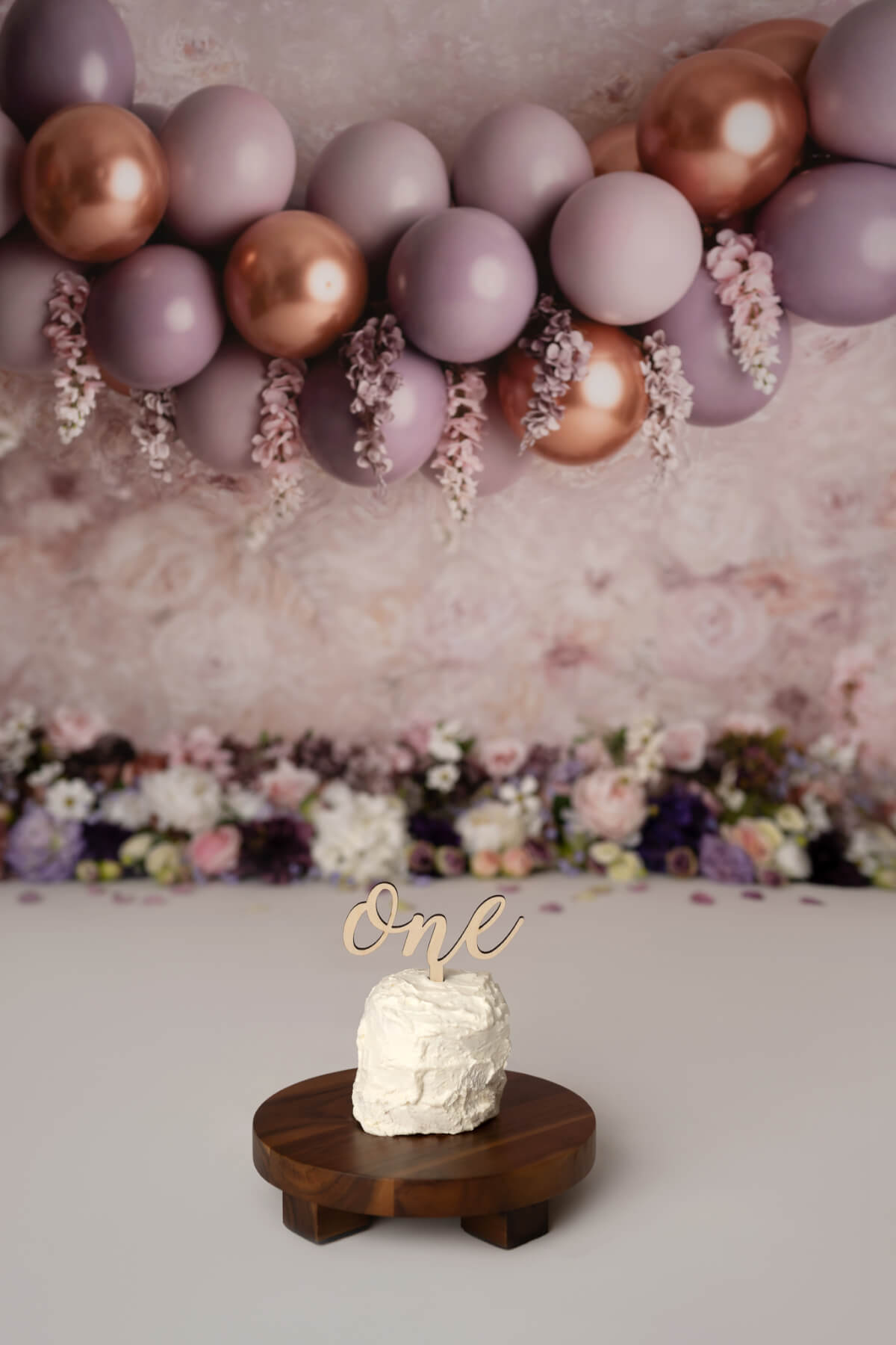 A small white cake with a topper that says "one" sits on a cake stand in front of a floral background decorated with balloons
