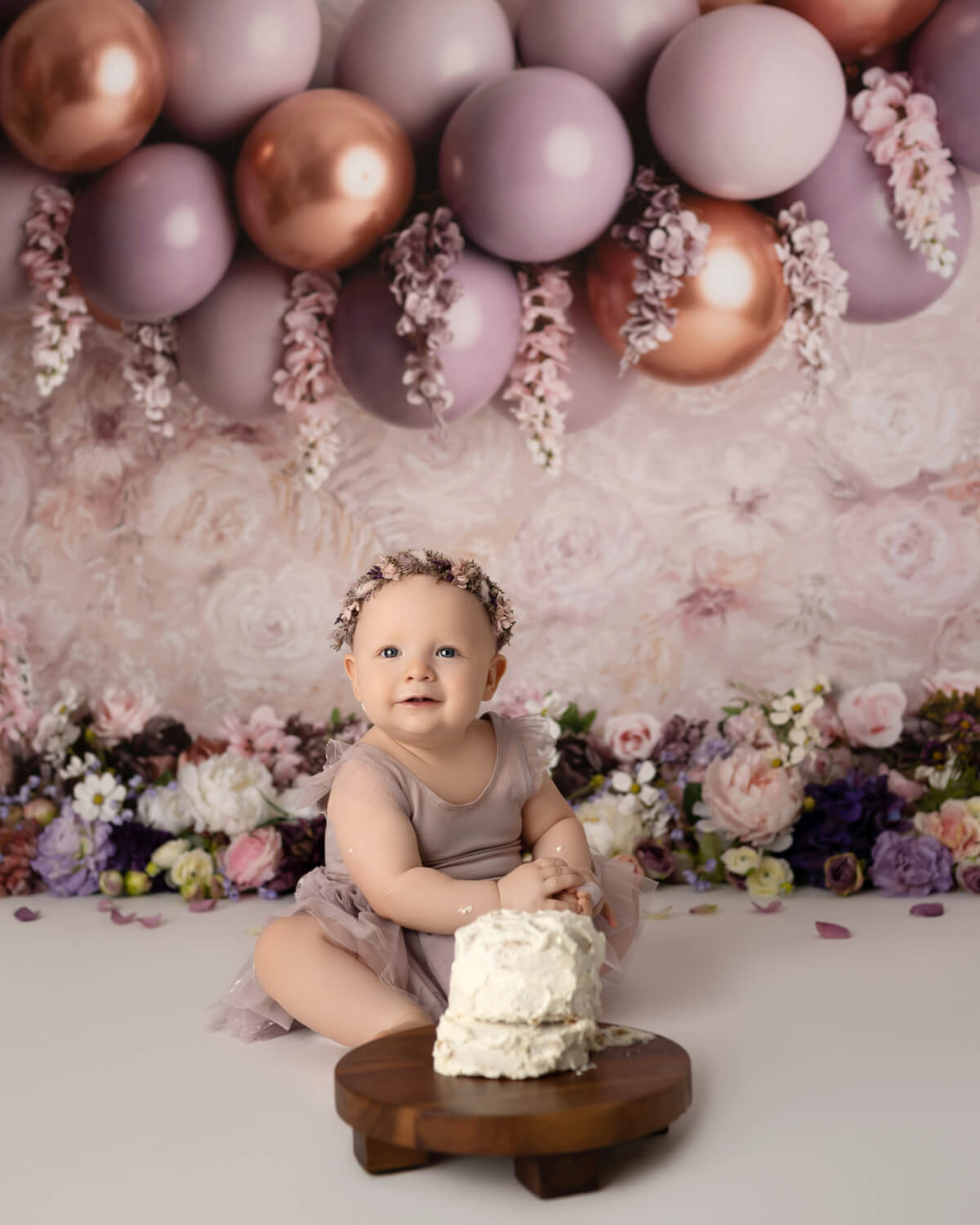 A baby sits in front of a floral background and balloons. There is a smashed cake in front of her, and she has frosting on her arms. 