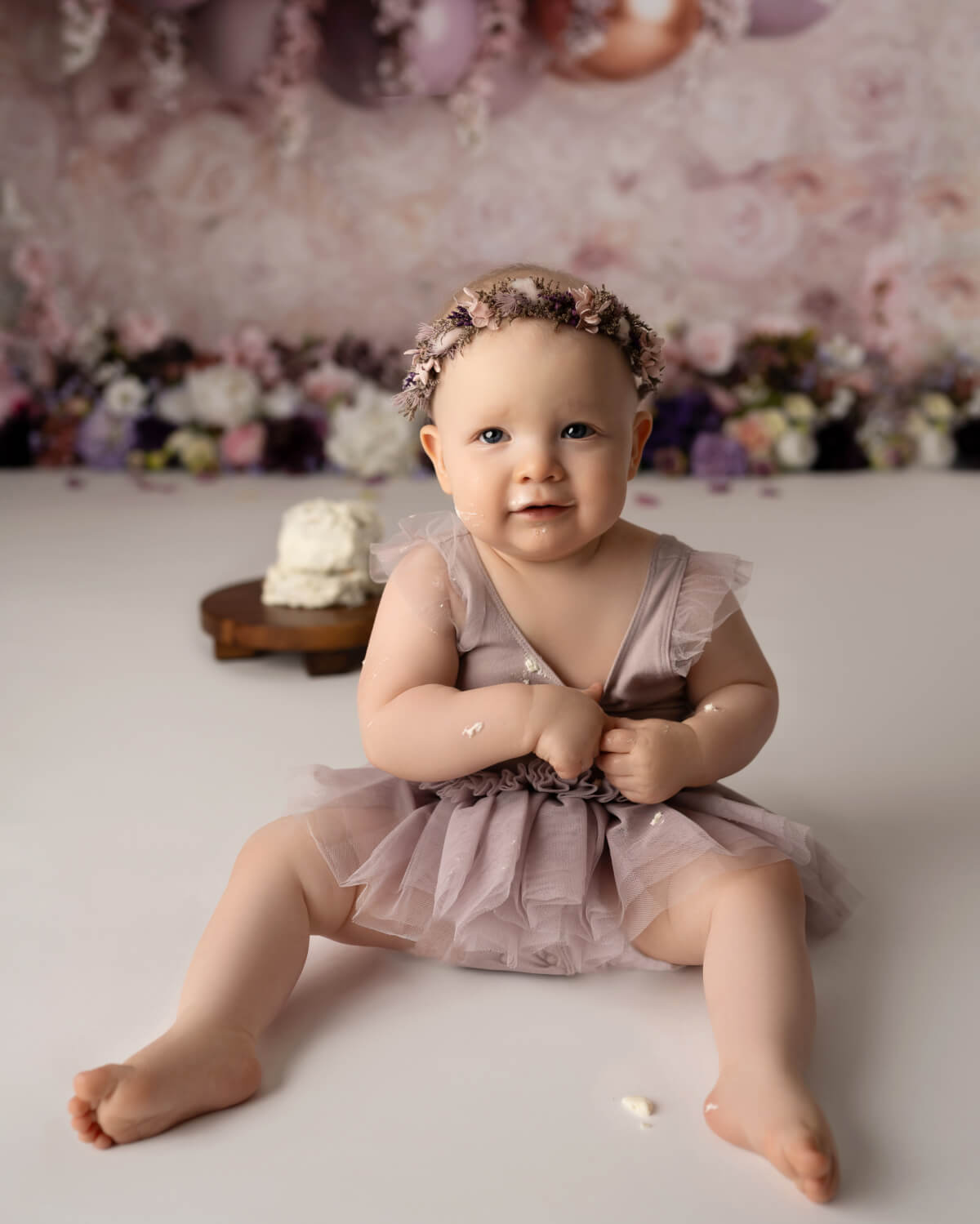 a baby sits in front of a smashed cake and a floral background. The baby is wearing a floral headband and lavender dress.