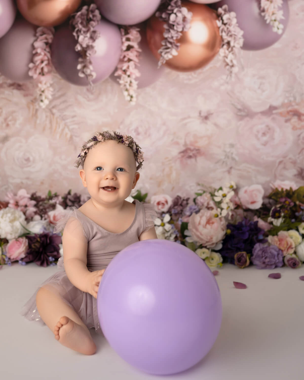 A baby in a lavender dress sits in front of balloons and flowers. She is holding a purple balloon. 