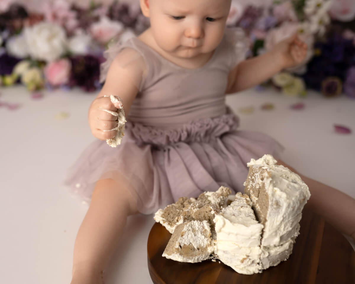 A baby in a lavender dress stares at a smashed white cake. The baby is holding a piece of cake in its hand.