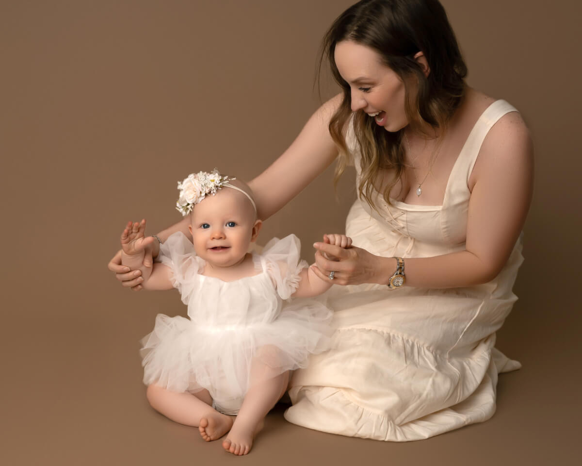 A woman in a white dress sits next to a baby in a white dress. They are both smiling, and the woman is holding up the baby's arms. 