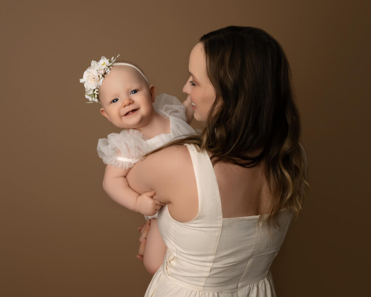 A woman in a white dress holds a baby in a white dress. The baby is smiling at the camera over the woman's shoulder.