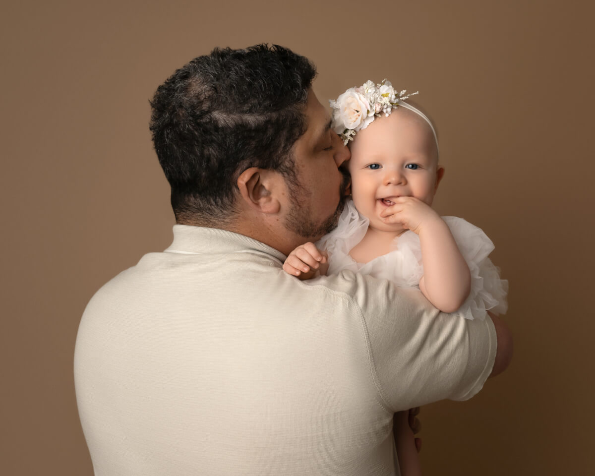 A man holds a baby in a white dress. The baby is looking over the man's shoulder and has her hand in her mouth.