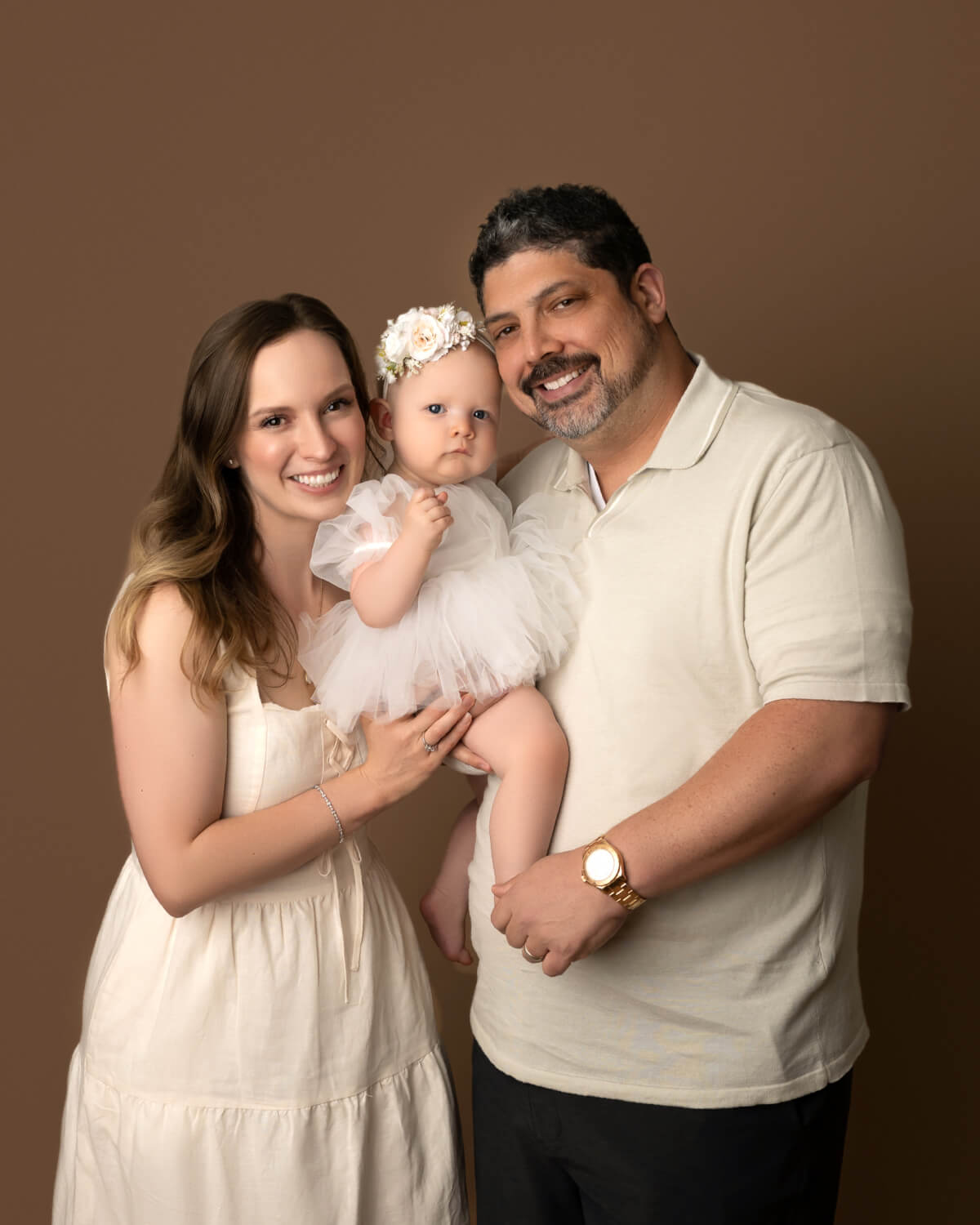 A man and a woman in white hold a baby in a white dress between them.