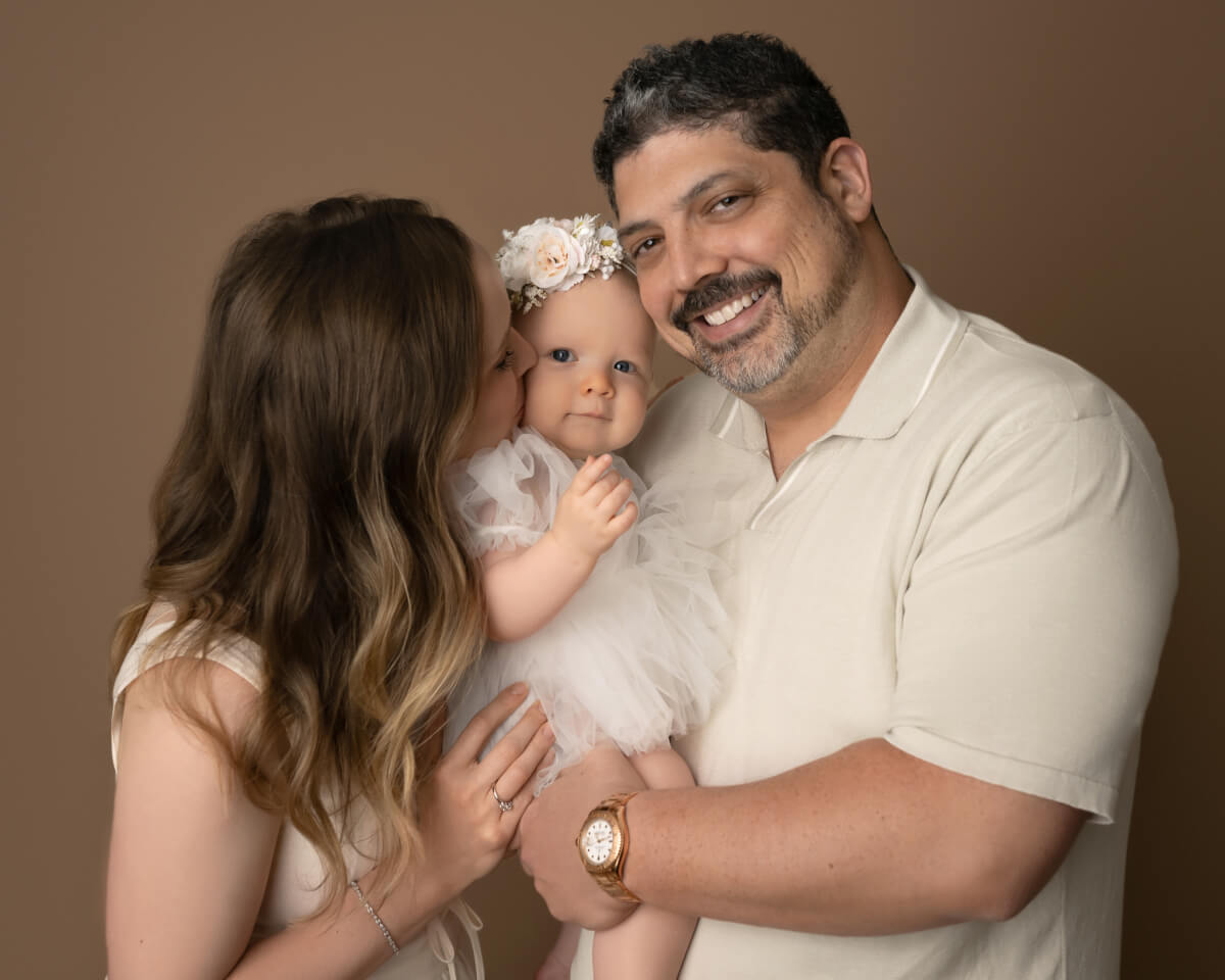 A man and a woman in white hold a baby in a white dress between them. The woman is kissing the baby on the cheek.