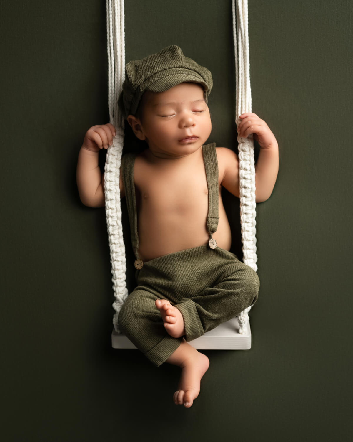 Newborn boy in green corduroy outfit on suspended swing set against a dark olive backdrop | Studio Newborn Photography Austin