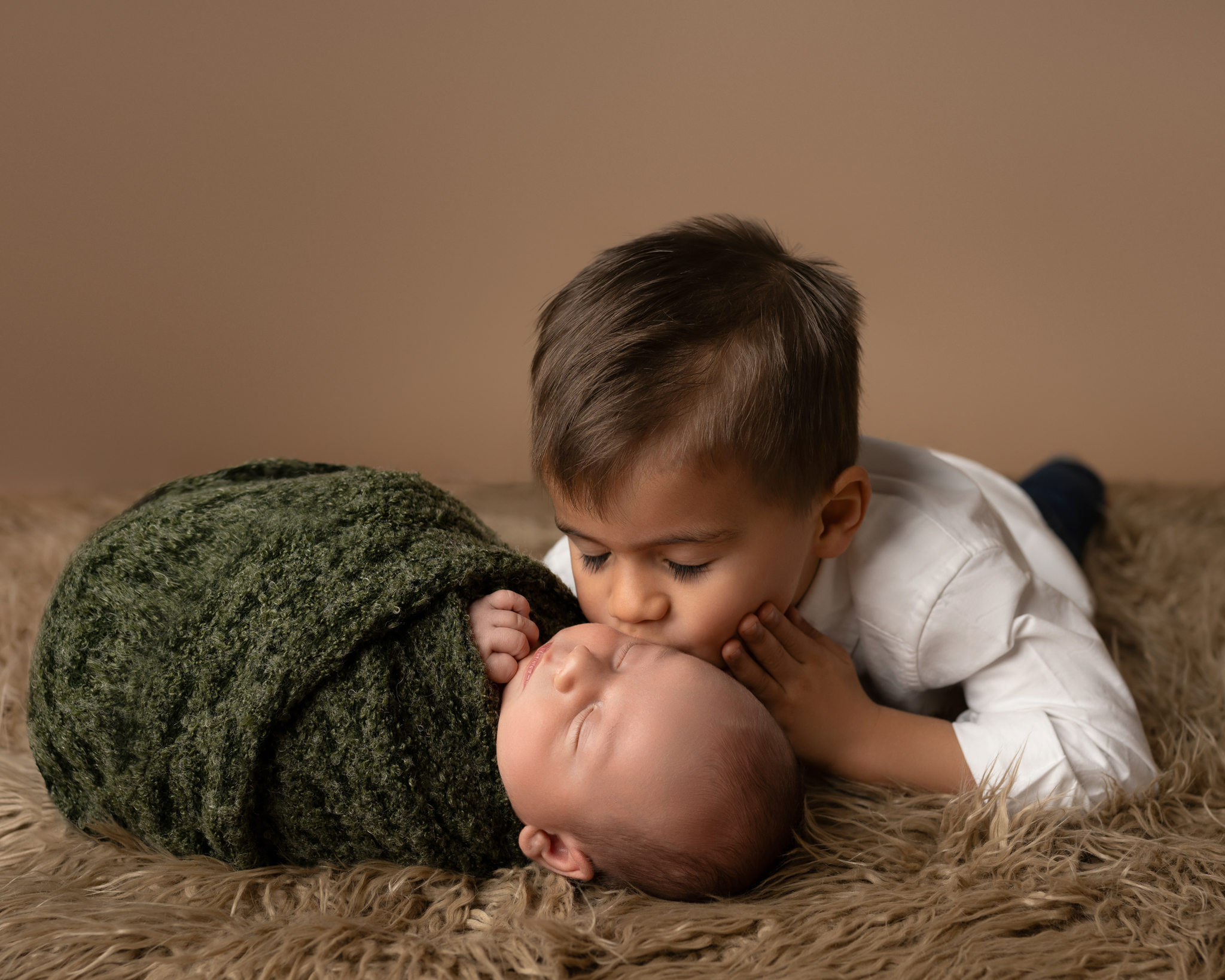 Older brother kissing newborn wrapped in deep green on faux fur blanket | Heartwarming sibling photo in Austin, TX