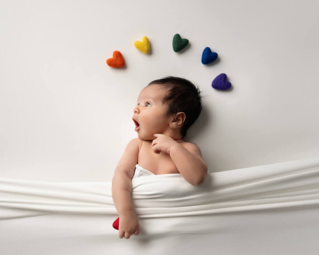 baby boy with rainbow hearts above his head tucked in a white bed to symbolize rainbow baby