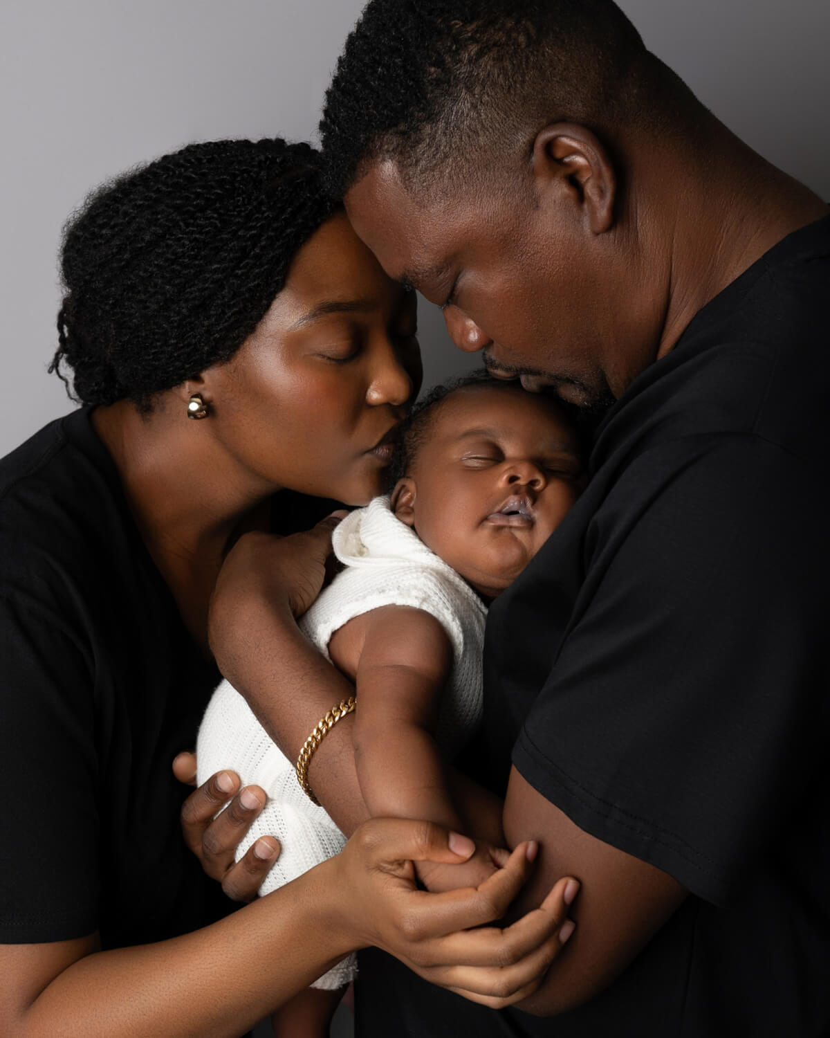 A man and a woman hold a sleeping newborn baby between them. They are both kissing the baby's head.
