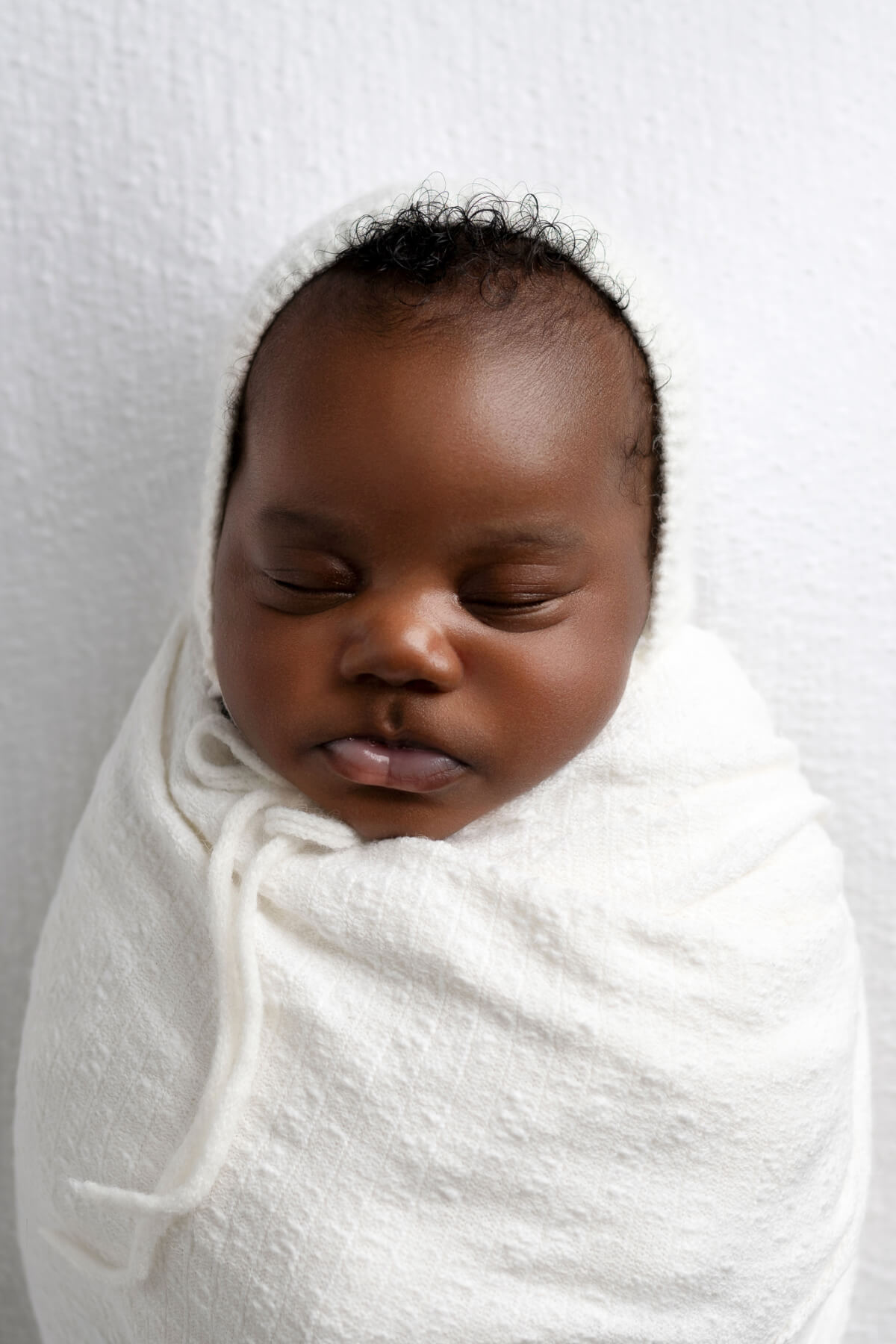 A color photo of a sleeping newborn baby swaddled in a white blanket. 