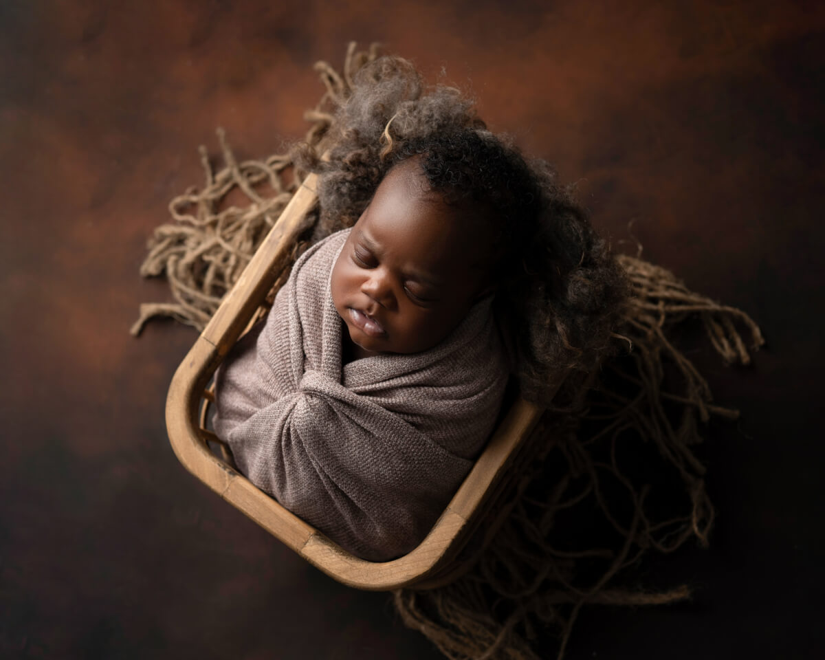 A newborn photo of a baby swaddled in a brown cloth and placed in a prop basket.