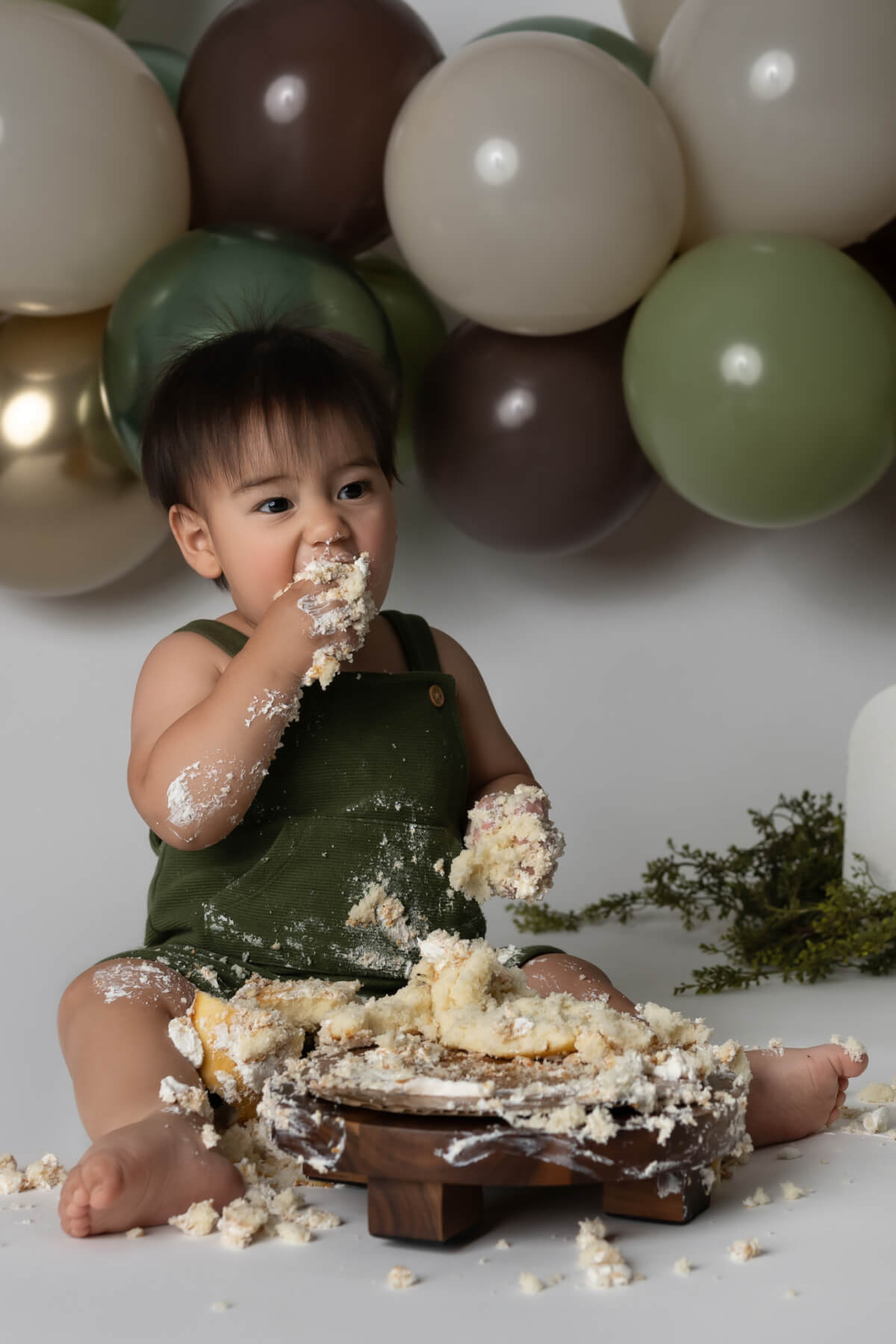 Diego at his first birthday cake smash session in Austin, wearing a green romper and enjoying cake with a balloon garland in the background.