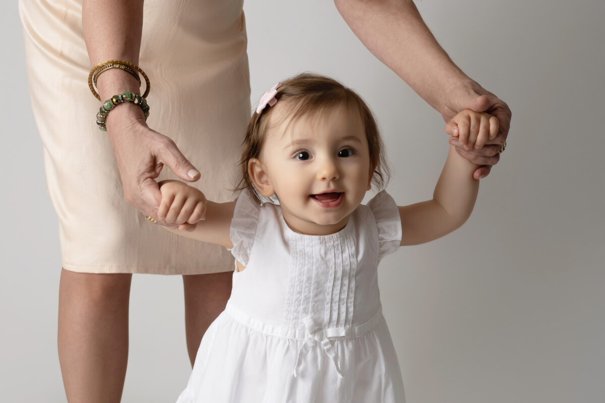 A baby in a white dress stands with the help of a woman. The woman stands behind her holding her hands. 