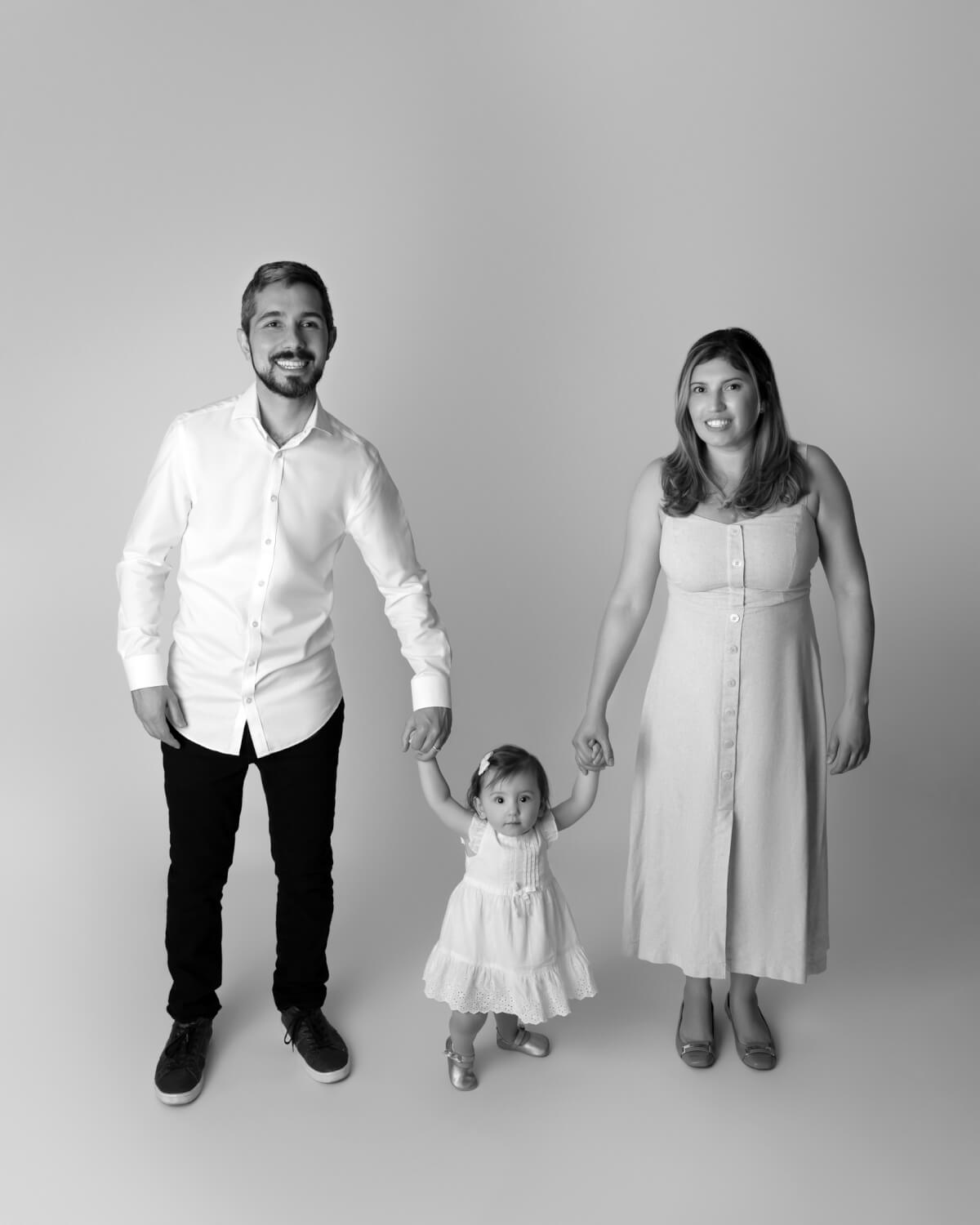 A black and white photo of a man, woman, and one year old baby. The baby is standing in the middle of the photo holding her parent's hands. 