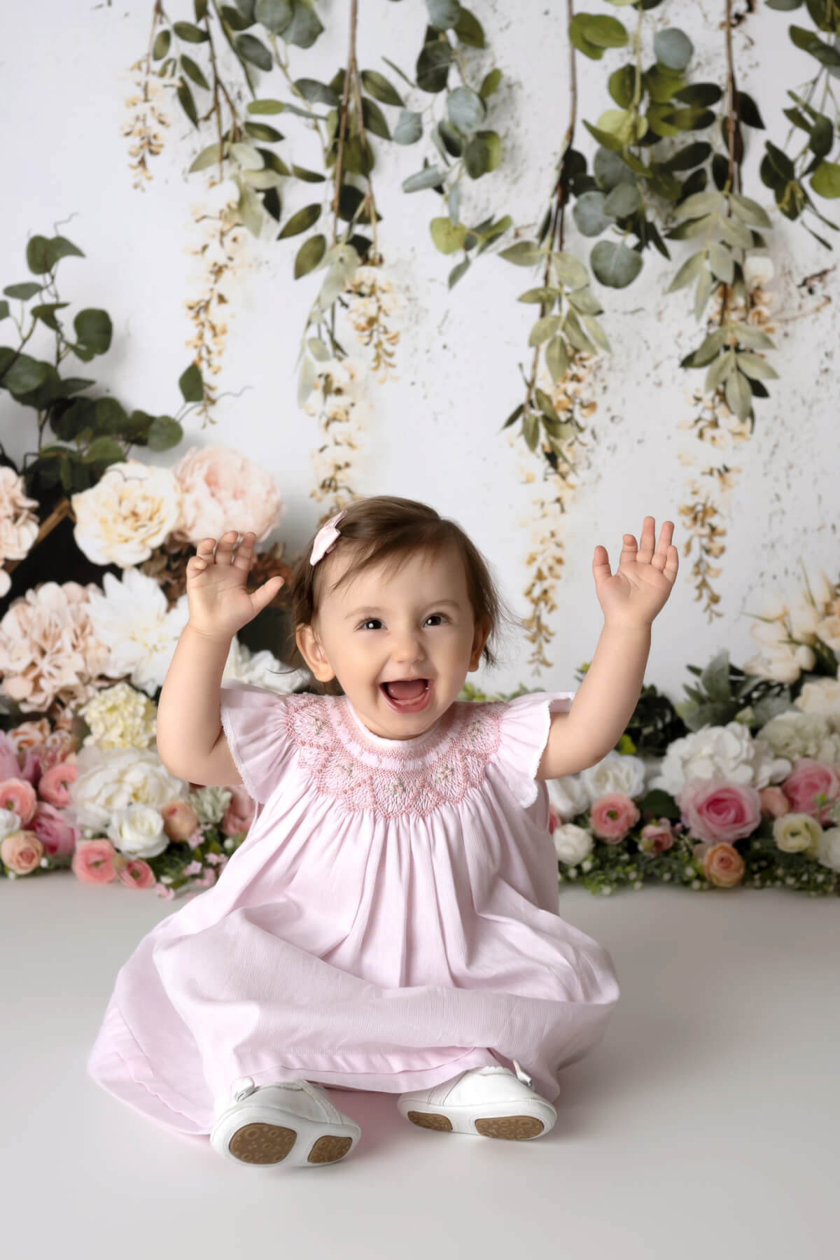 A one year old girl sits in front of a flower garland in a photo studio. She is laughing and has her hands raised in the air.
