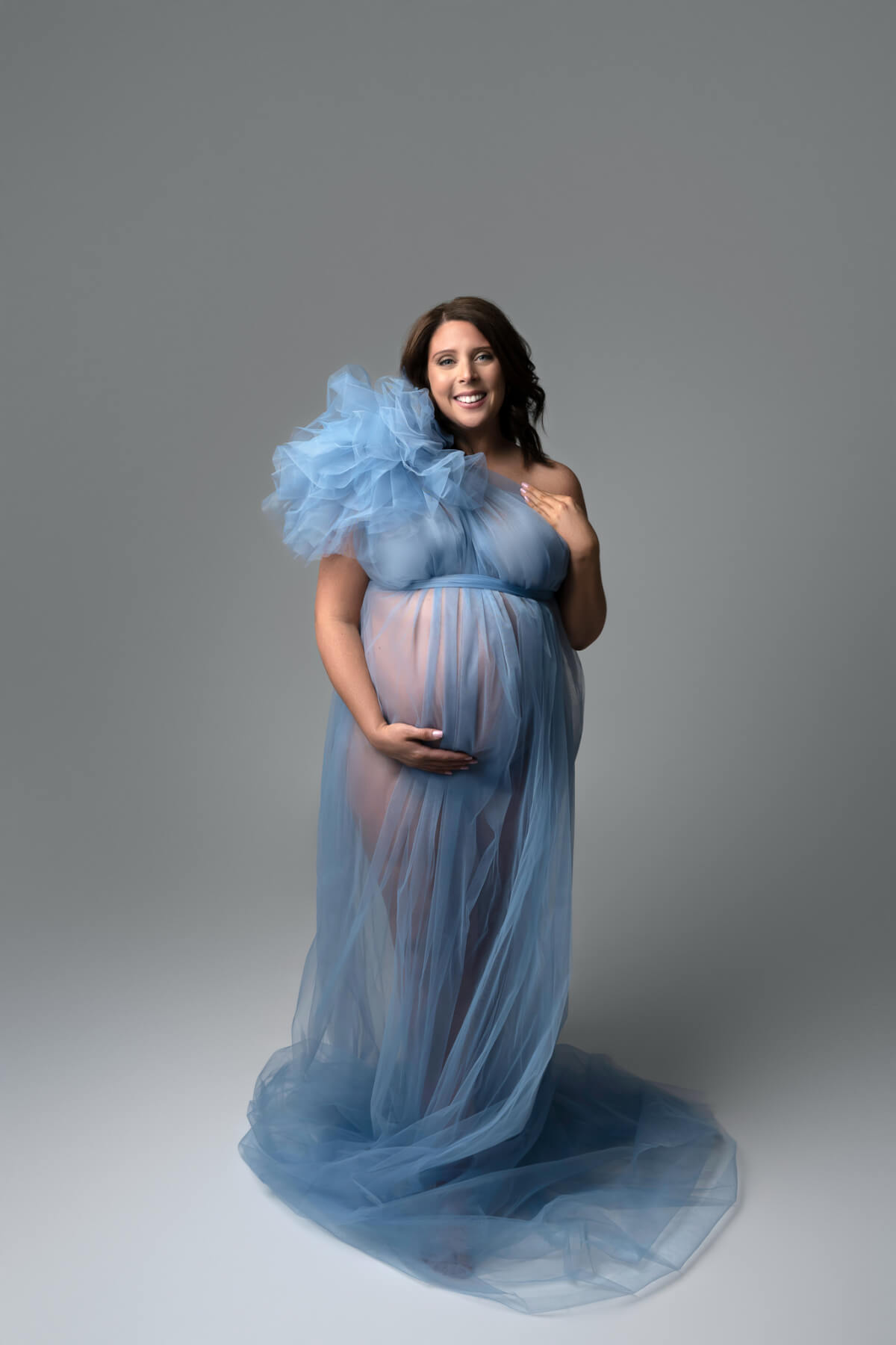 A pregnant woman in a dusky blue tulle dress holds her baby bump. She is smiling and looking straight into the camera.