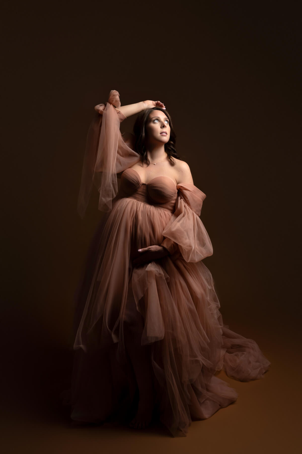 A pregnant woman faces a bright light over her left shoulder. She is holding one hand on the underside of her belly and the other on the top of her head. 