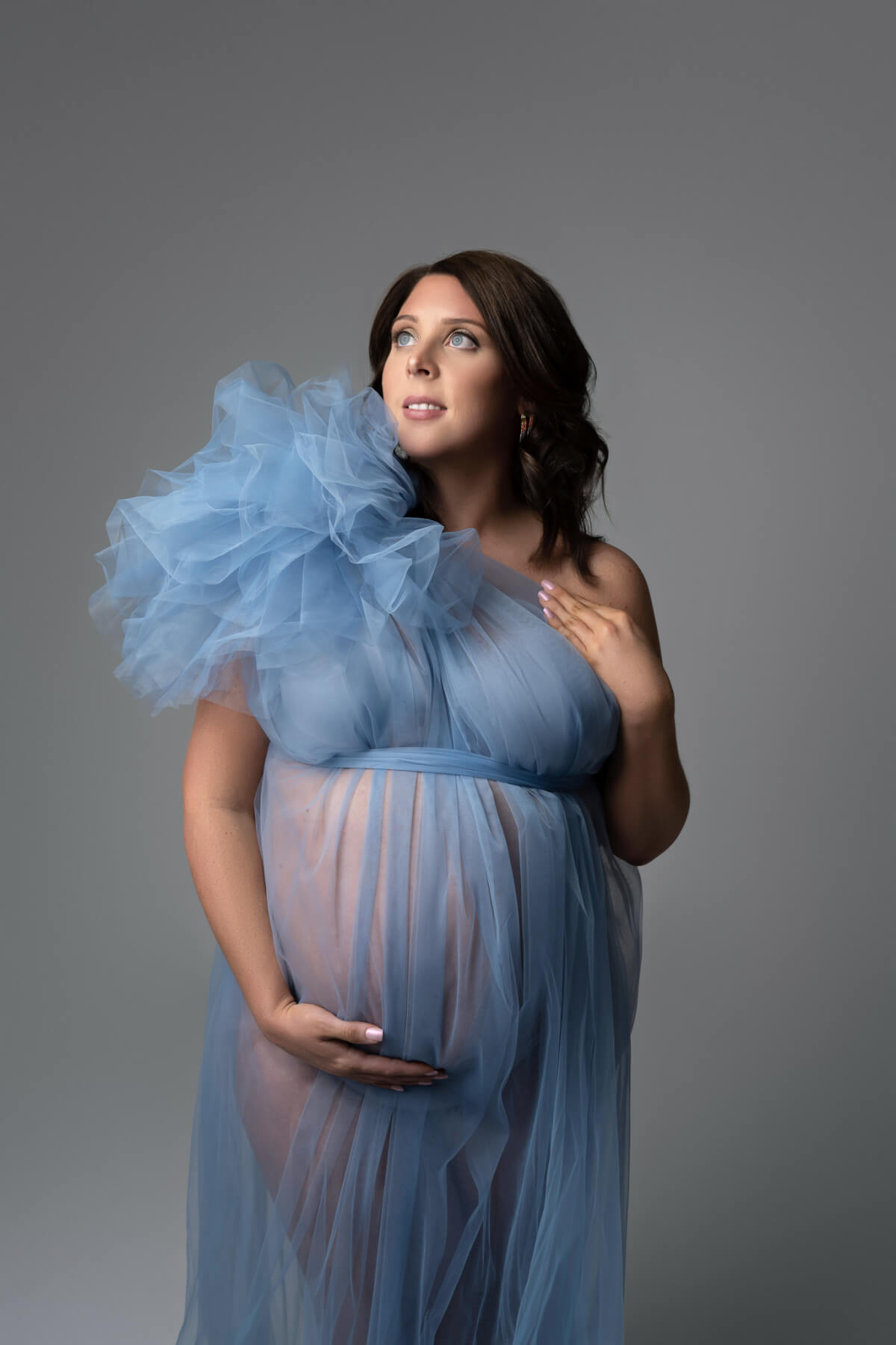 A pregnant woman in a dusky blue tulle gown looks off to her right. She is holding the underside of her belly. 