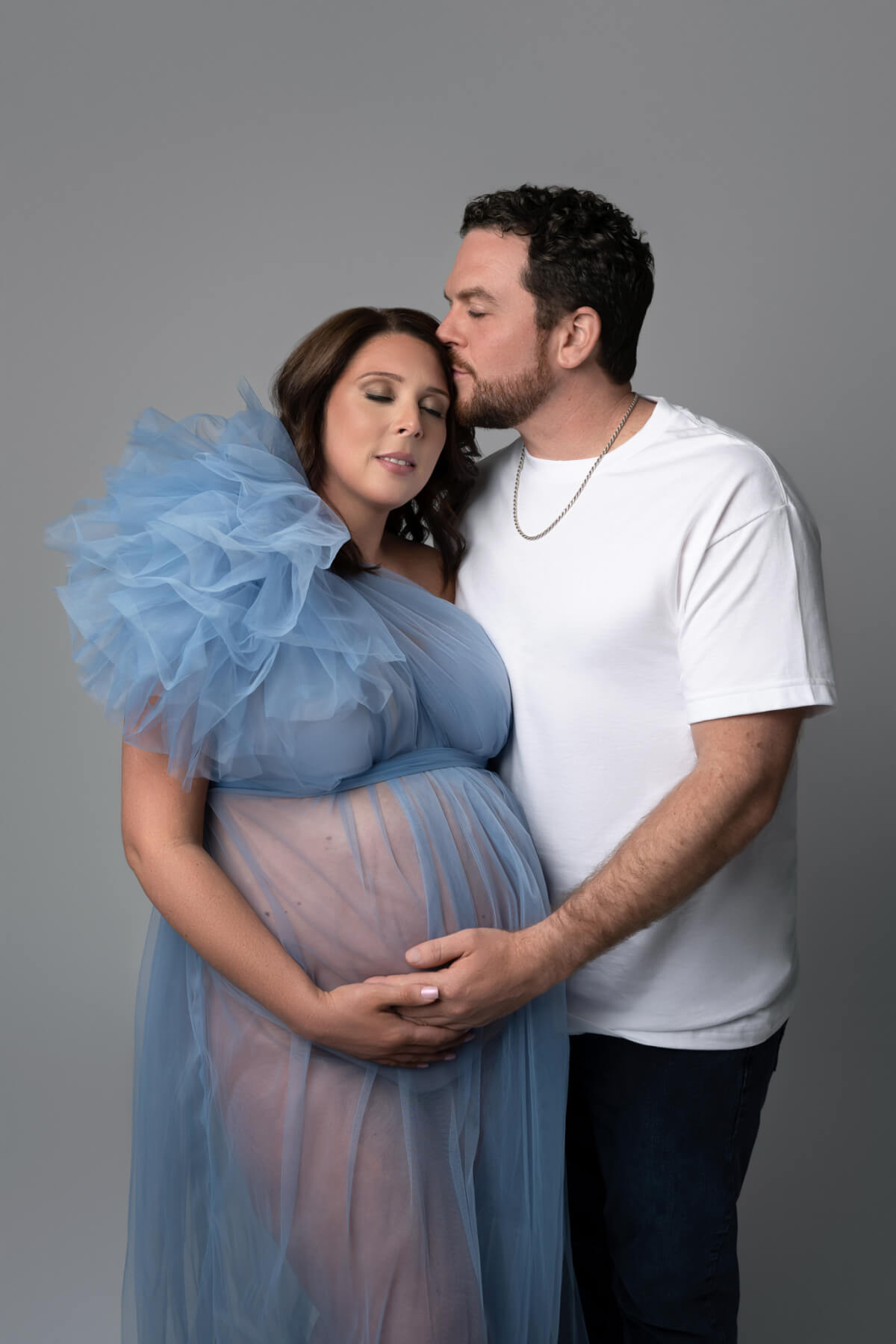 A man and a woman stand side-by-side in a Cedar Park photography studio. The man is kissing the woman's forehead and they both have a hand on the underside of her belly. 