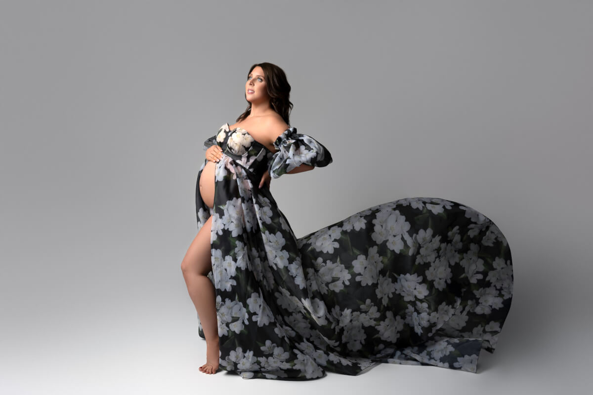 A pregnant woman in a Cedar Park maternity studio stands in side profile with one hand on the top of her belly and one on her back. She is wearing a black floral maternity gown.