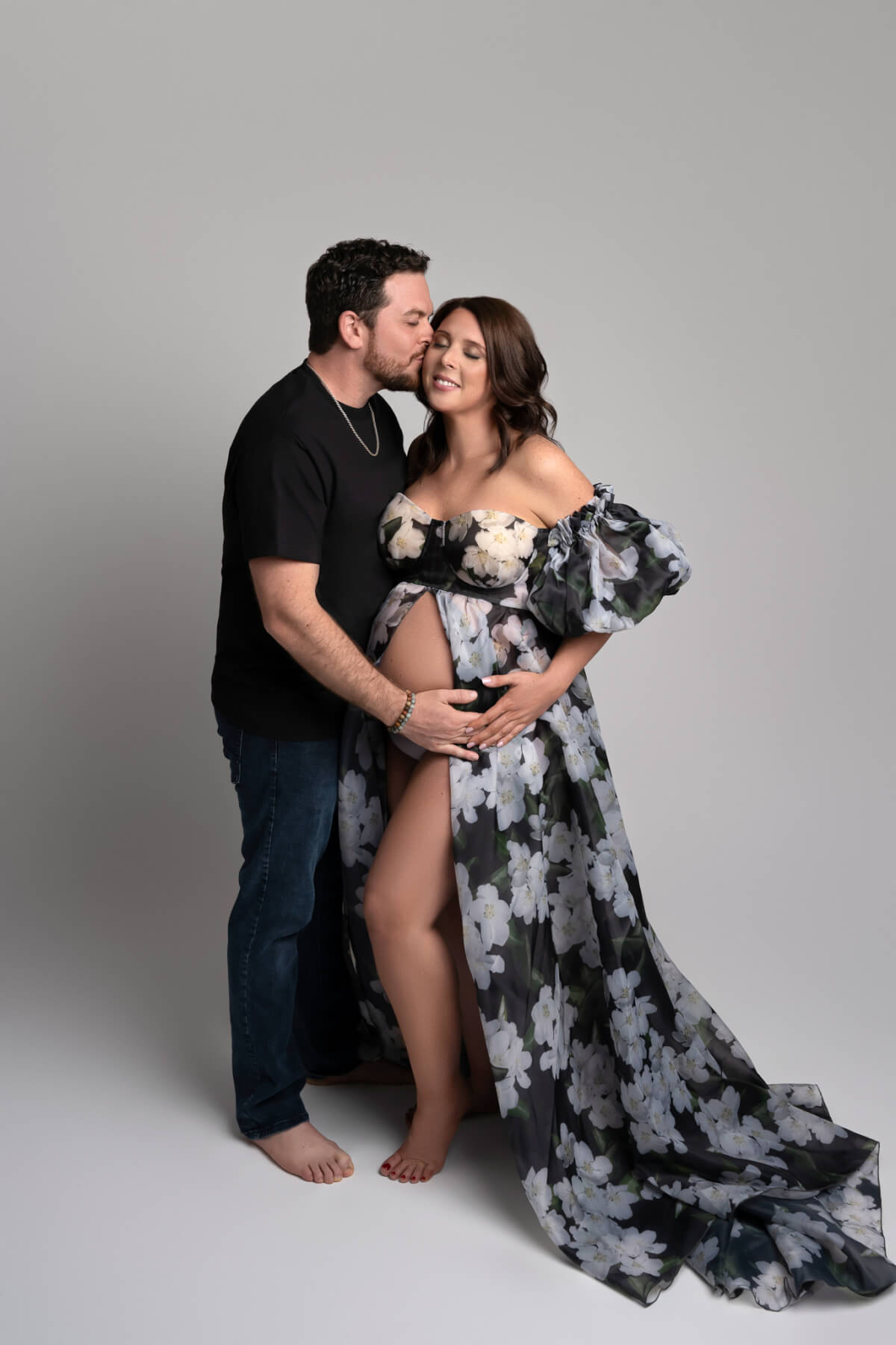 A man kisses a pregnant woman on the cheek. They both have one hand on her belly.