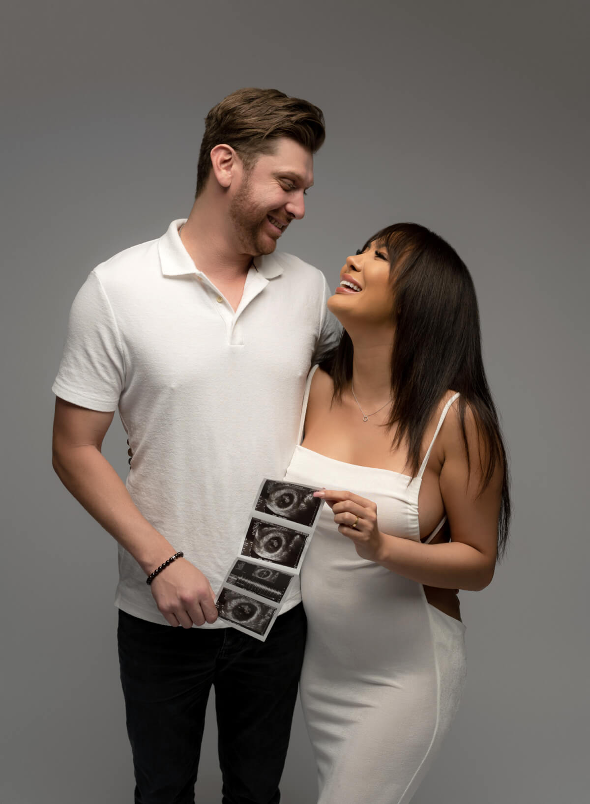 A woman in a white dress and a man in a white polo stand next to each other. They are smiling and looking into one another's eyes and holding a set of ultrasound photos in front of them.