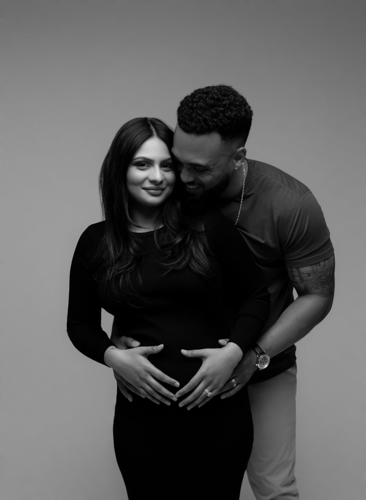 A man and a woman in a modern photography studio. They both have their hands on her baby bump. 