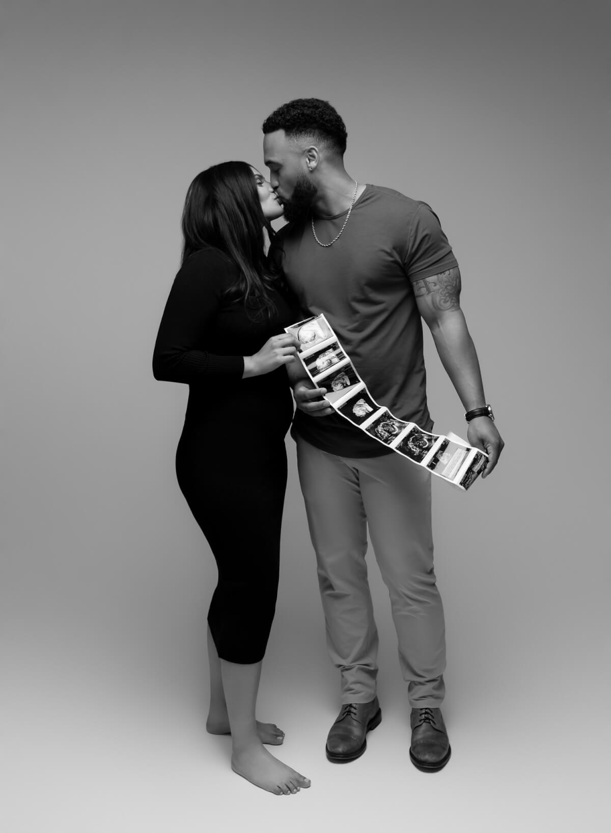 A man and a woman holding a string of ultrasound photos kiss each other. 