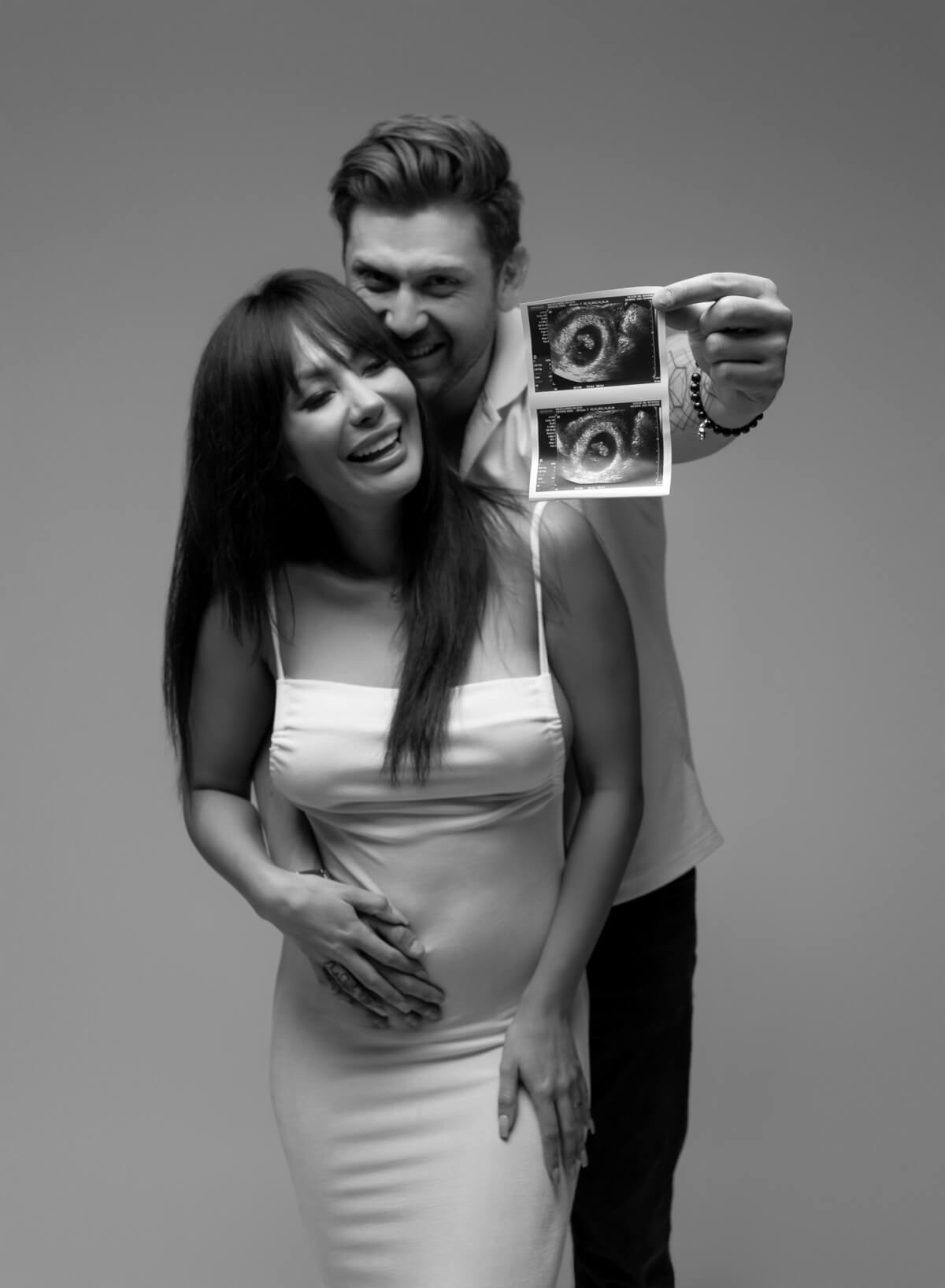 A smiling man stands beside a smiling woman. He has one hand on hers, which is placed on her stomach, and his other hand is holding up a sonogram photo. 