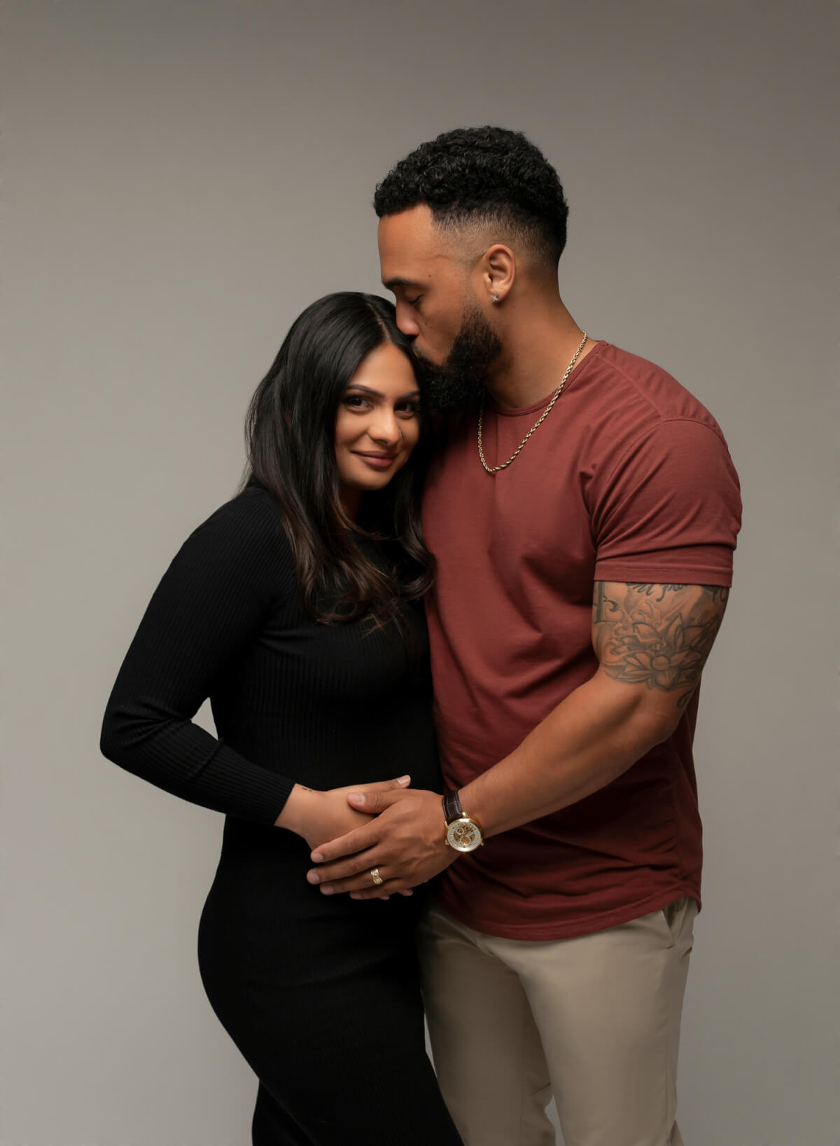 A man and woman in a modern pregnancy photography studio stands next to one another. The man is kissing the woman on the forehead, and both have one hand on the woman's stomach.