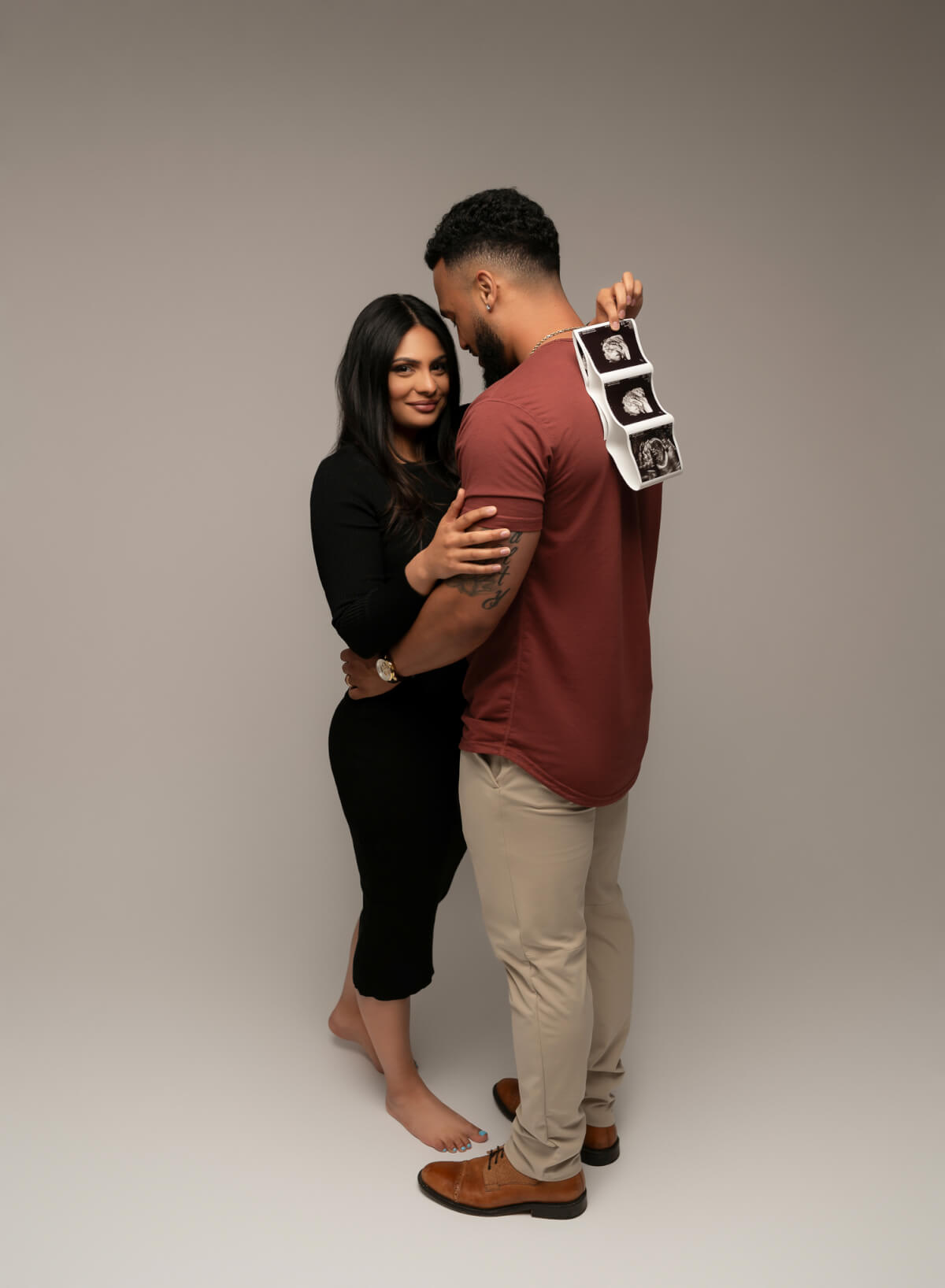 A man and woman stand on a photography studio set facing one another. The woman is holding a string of ultrasound photos over the man's shoulder.