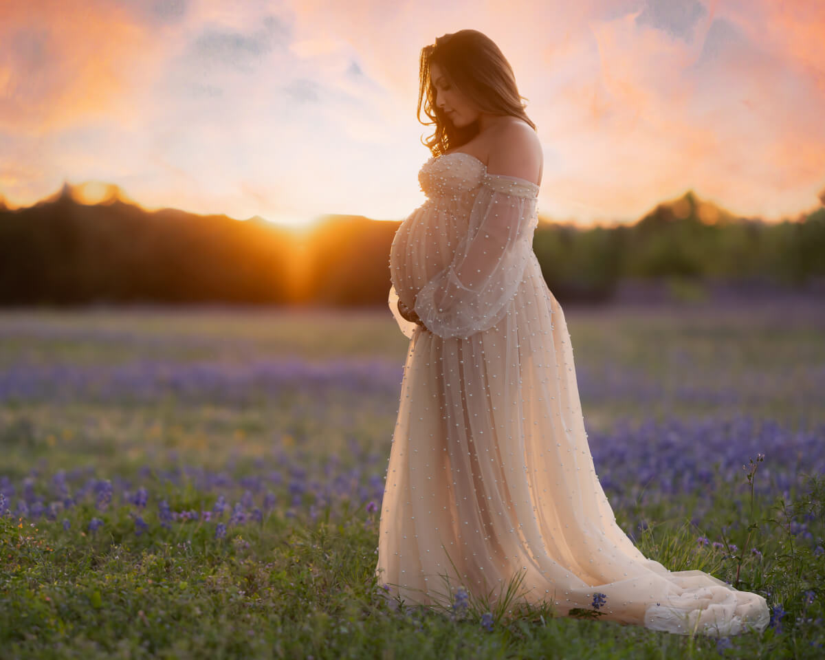 pregnancy photoshoot mom wearing ivory pearl gown standing in a field of bluebonnets with dramatic sky