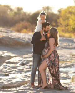 dad kissing pregnant mom with toddler on his shoulders covering her eyes