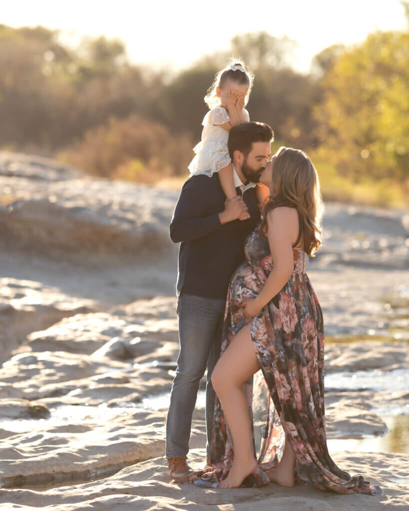 dad kissing pregnant mom with toddler on his shoulders covering her eyes