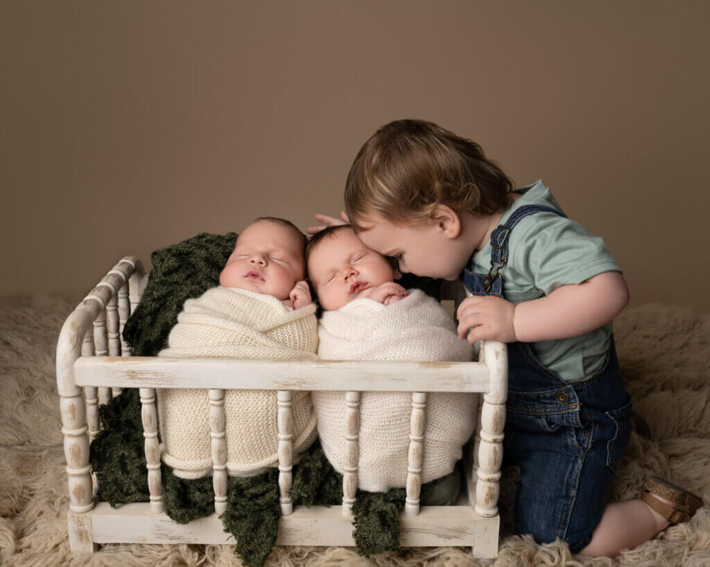 twin baby boys wrapped in white placed in tiny crib with toddler sibling touching foreheads