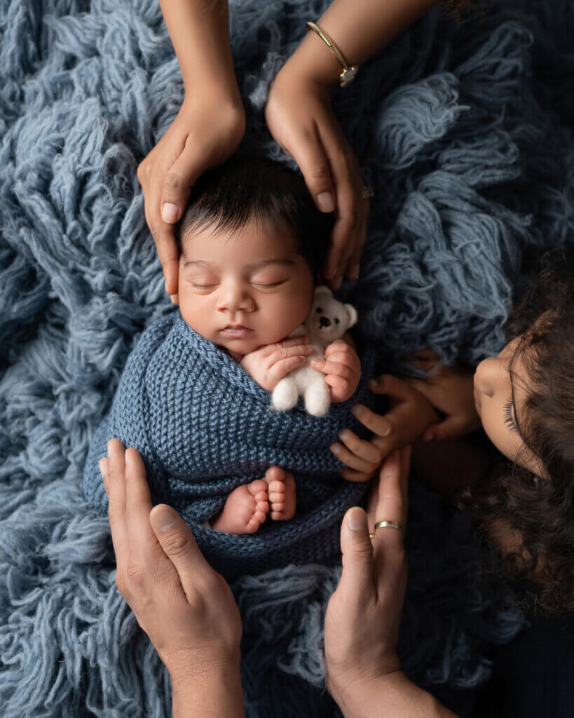 Baby wrapped in blue knit laying on blue rug with parents and big sister's hands surrounding baby boy