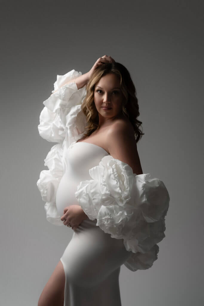 Confident pregnant woman in a white gown with voluminous ruffled sleeves looking directly at the camera.