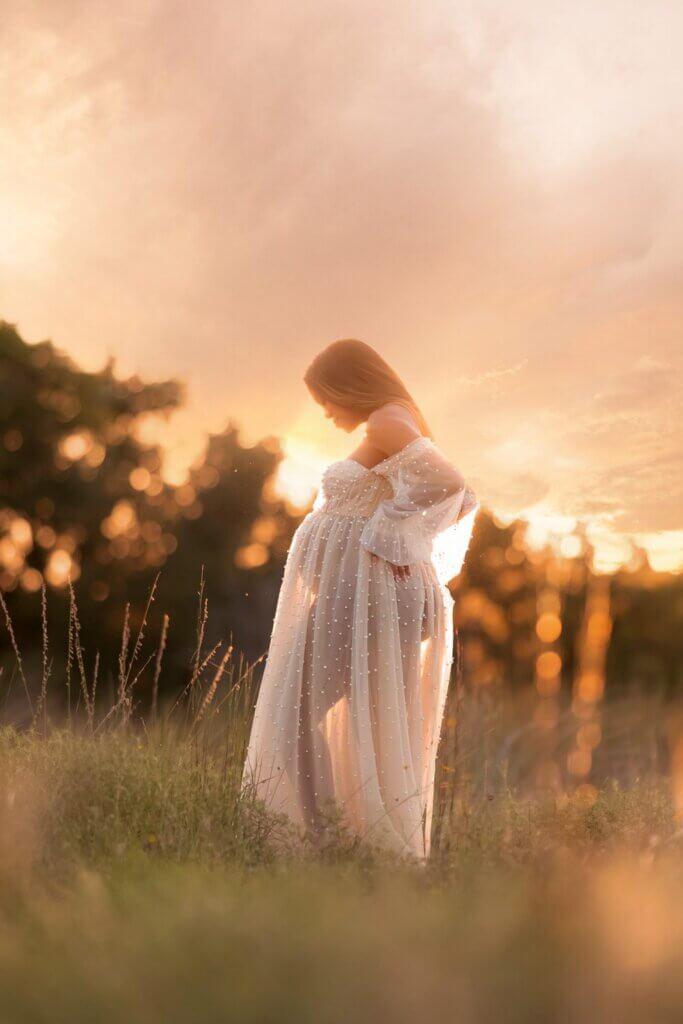 Backlit pregnant mother in a flowing white gown in a Hill Country field during a babymoon photo session